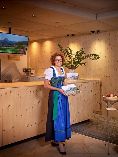 Een vrouw in traditionele klederdracht staat bij de receptie van een houten hotel. De sfeer is vriendelijk en uitnodigend.