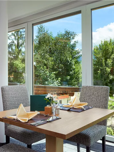 Een uitnodigend restaurant met uitzicht op groene bomen en bergen. De tafels zijn mooi gedekt en de sfeer is vriendelijk en licht.