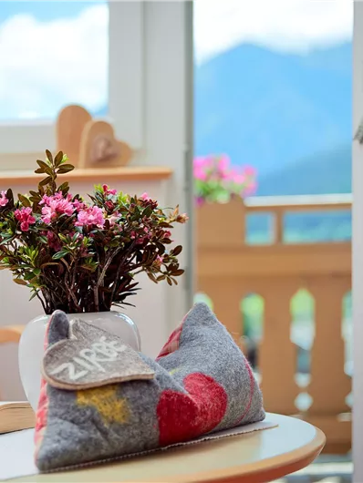 Een gezellige kamer met een kleine tafel, waarop een bloem en een boek liggen. Het uitzicht gaat door het raam op de bergen en het balkon.
