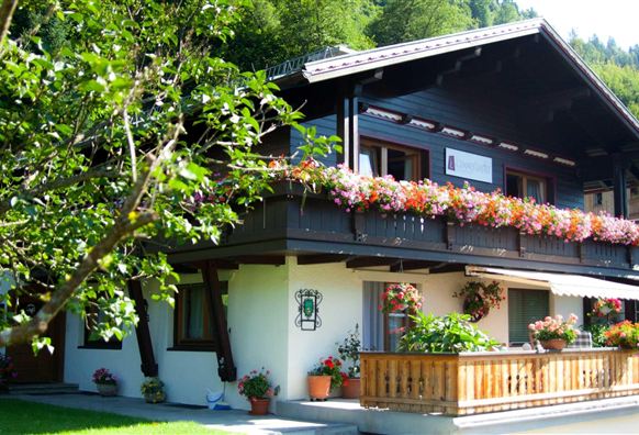 A cozy wooden house with a balcony full of blooming flowers. The surroundings are green and inviting.