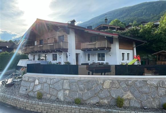 A modern house with wooden cladding and balconies, surrounded by greenery. In the foreground, a paved driveway and a stone wall can be seen.