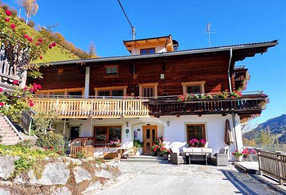 A traditional wooden house in the mountains with a large balcony and flowers. In the background, green hills and a blue sky are visible.