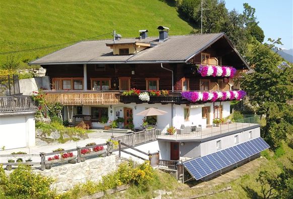 Ein charmantes Holzhaus mit großen Balkonen, geschmückt mit bunten Blumen. Im Vordergrund sind ein Garten und Solarpanels zu sehen, umgeben von grünen Hügeln.