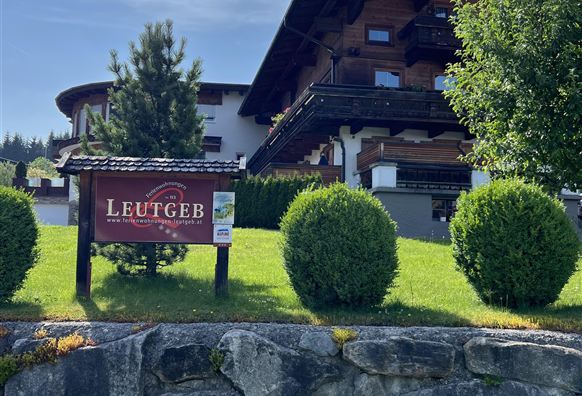 A traditional Tyrolean chalet surrounded by well-kept greenery. In the foreground, there is a sign with the inscription "Leutgeb".