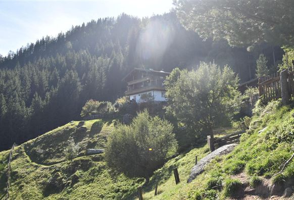 Ein malerisches Haus auf einem grünen Hügel, umgeben von Bäumen. Im Hintergrund erheben sich hohe Berge unter einem klaren Himmel.