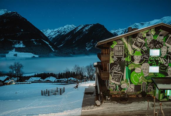 Ein modernes altes Holzhaus mit einer bunten Wandgestaltung. Im Hintergrund sind schneebedeckte Berge und ein nebliges Tal zu sehen.