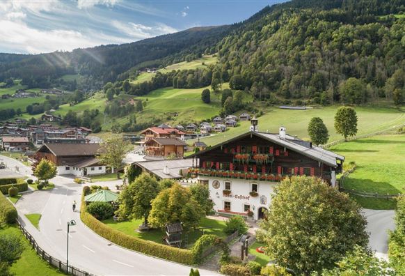 Ein malerisches Alpenpanorama mit grünen Wiesen und Bergen. Im Vordergrund steht ein traditionelles Gebäude, umgeben von Bäumen und einer ruhigen Landschaft.