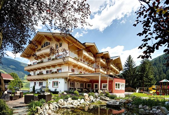 Ein gemütliches Hotel mit einem traditionellen österreichischen Architekturstil, umgeben von Bäumen und einer schönen Landschaft. Der Garten bietet eine entspannende Atmosphäre mit einem Teich und Spielplatz.