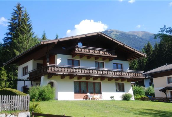 Ein traditionelles Haus in den Alpen mit einem Balkon und viel Grün im Vordergrund. Im Hintergrund sind Wälder und Berge sichtbar.