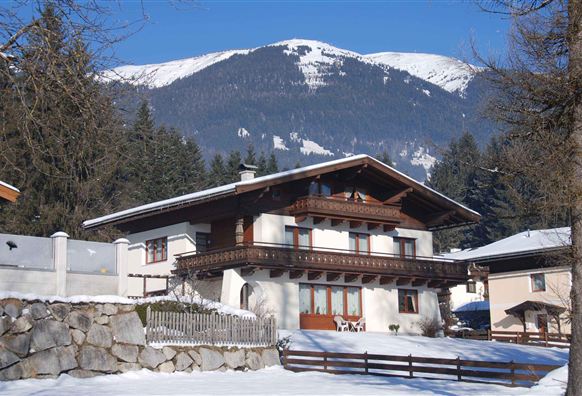Ein traditionelles Chalet in einer verschneiten Landschaft mit Bergen im Hintergrund. Die Umgebung ist von Bäumen umgeben und strahlt eine ruhige Winteratmosphäre aus.