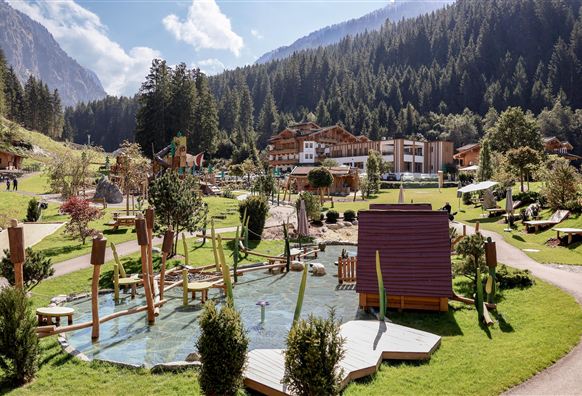 Ein liebevoll gestalteter Spielplatz mit Wasseranlagen und Spielgeräten. Im Hintergrund sind Wälder und Berge zu sehen, sowie ein gemütliches Gebäude.