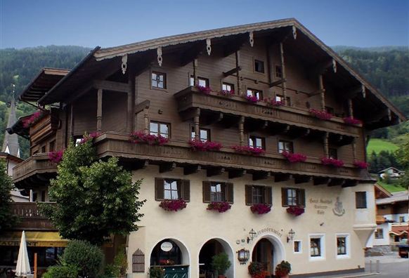 Ein traditionelles Holzhaus mit mehreren Balkonen und farbenfrohen Blumen. Im Hintergrund sind sanfte Hügel und Bäume zu sehen.