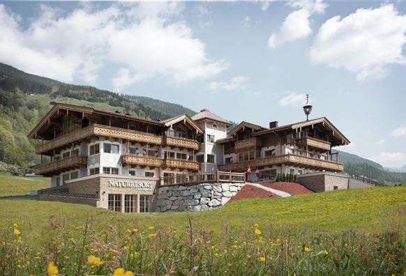 Ein charmantes Berghotel mit traditioneller Architektur inmitten einer grünen Wiese. Die Landschaft wird von sanften Hügeln und einem blauen Himmel umgeben.